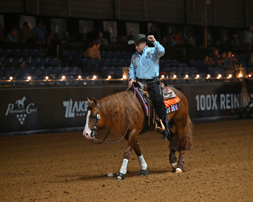 A rider raises a fist in celebration while sitting on a chestnut reining horse inside an arena, with spotlights and spectators in the background.