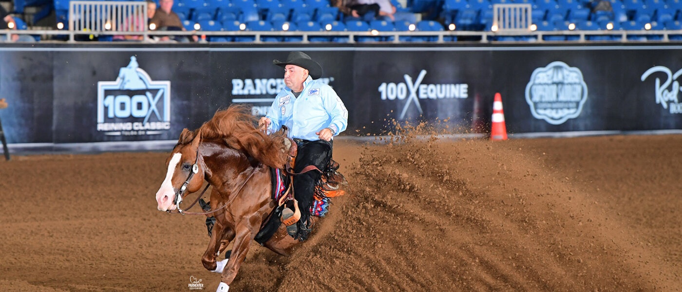 A reining horse and rider perform a sliding stop in an indoor arena, dirt flying as the horse digs into the ground with the crowd watching from the stands
