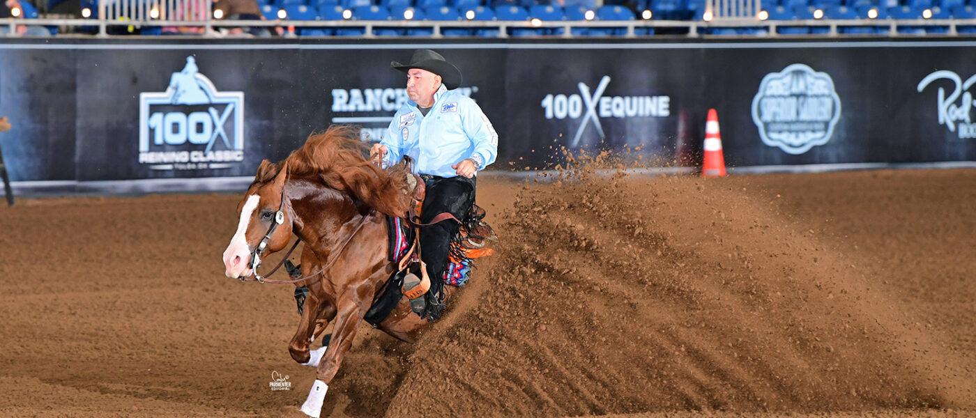 A reining horse and rider perform a sliding stop in an indoor arena, dirt flying as the horse digs into the ground with the crowd watching from the stands