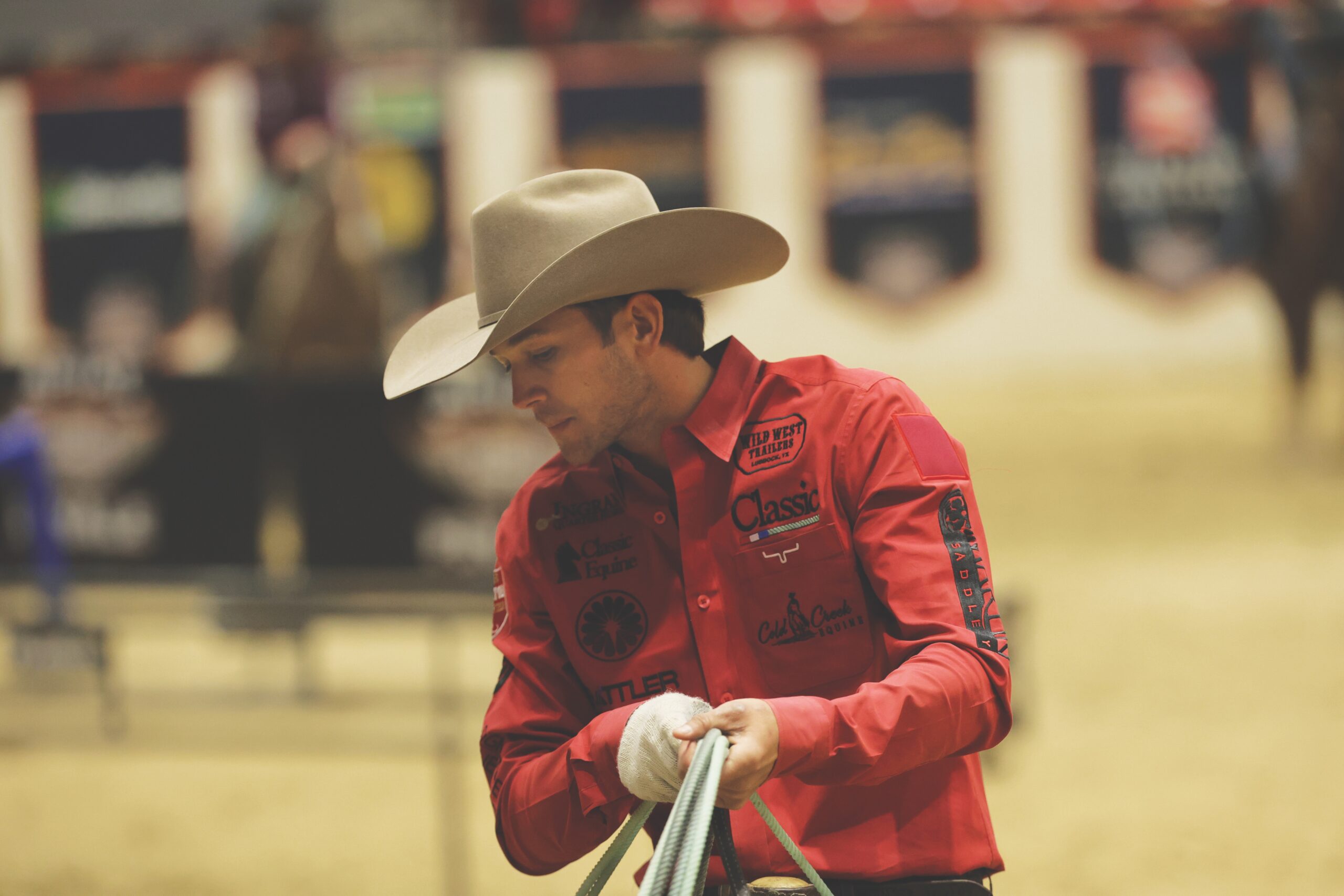 Team roper Paden Bray coils his rope in a red shirt and cowboy hat inside the arena