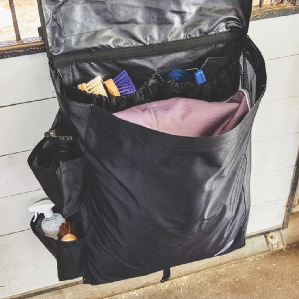 Black SmartPak blanket storage bag hanging on a stall front, holding a folded blanket and grooming brushes in its pockets.