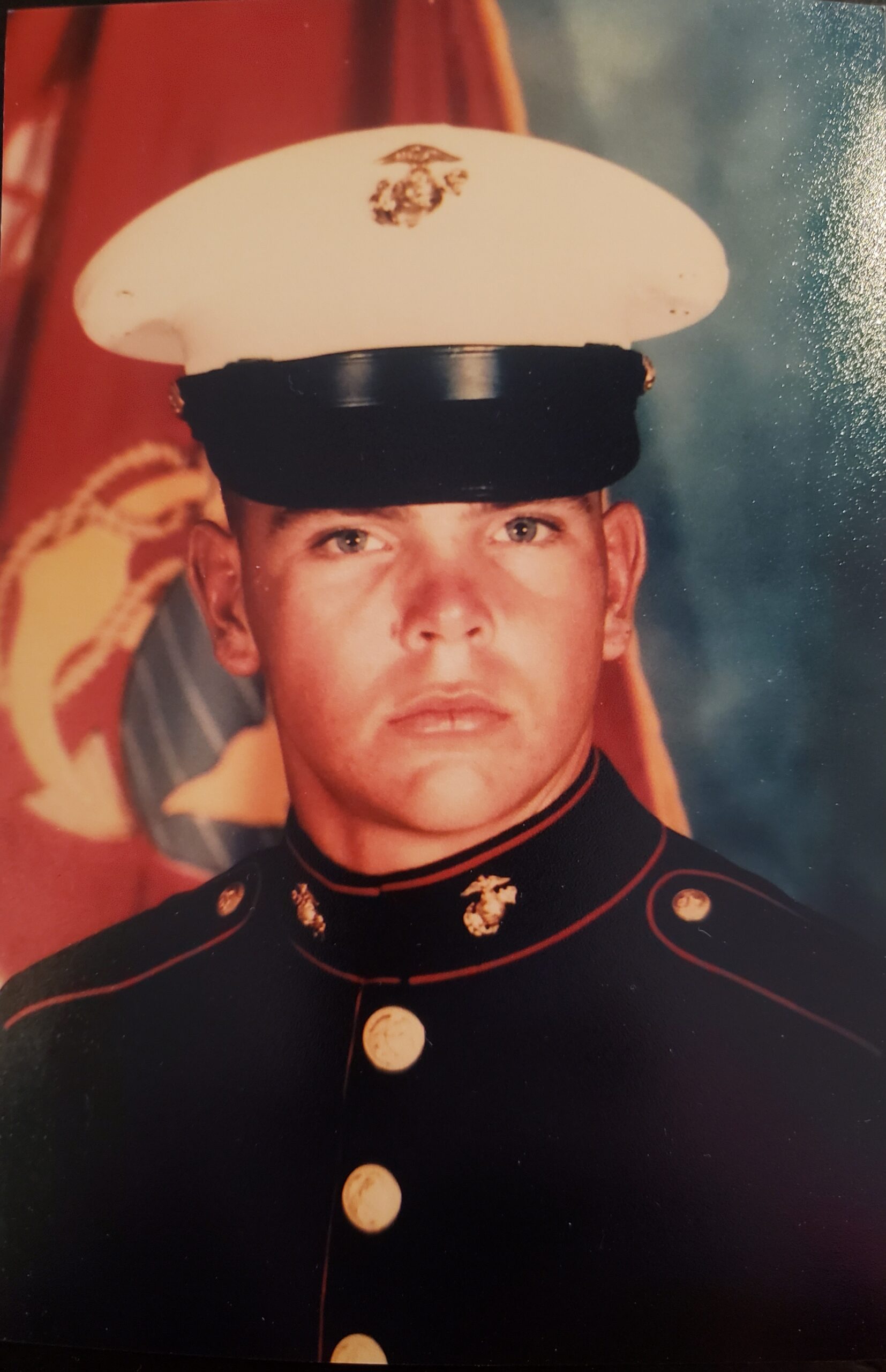 Bill Norman in Marine Corps dress blues and a white cover in front of a scarlet Marine Corps flag.