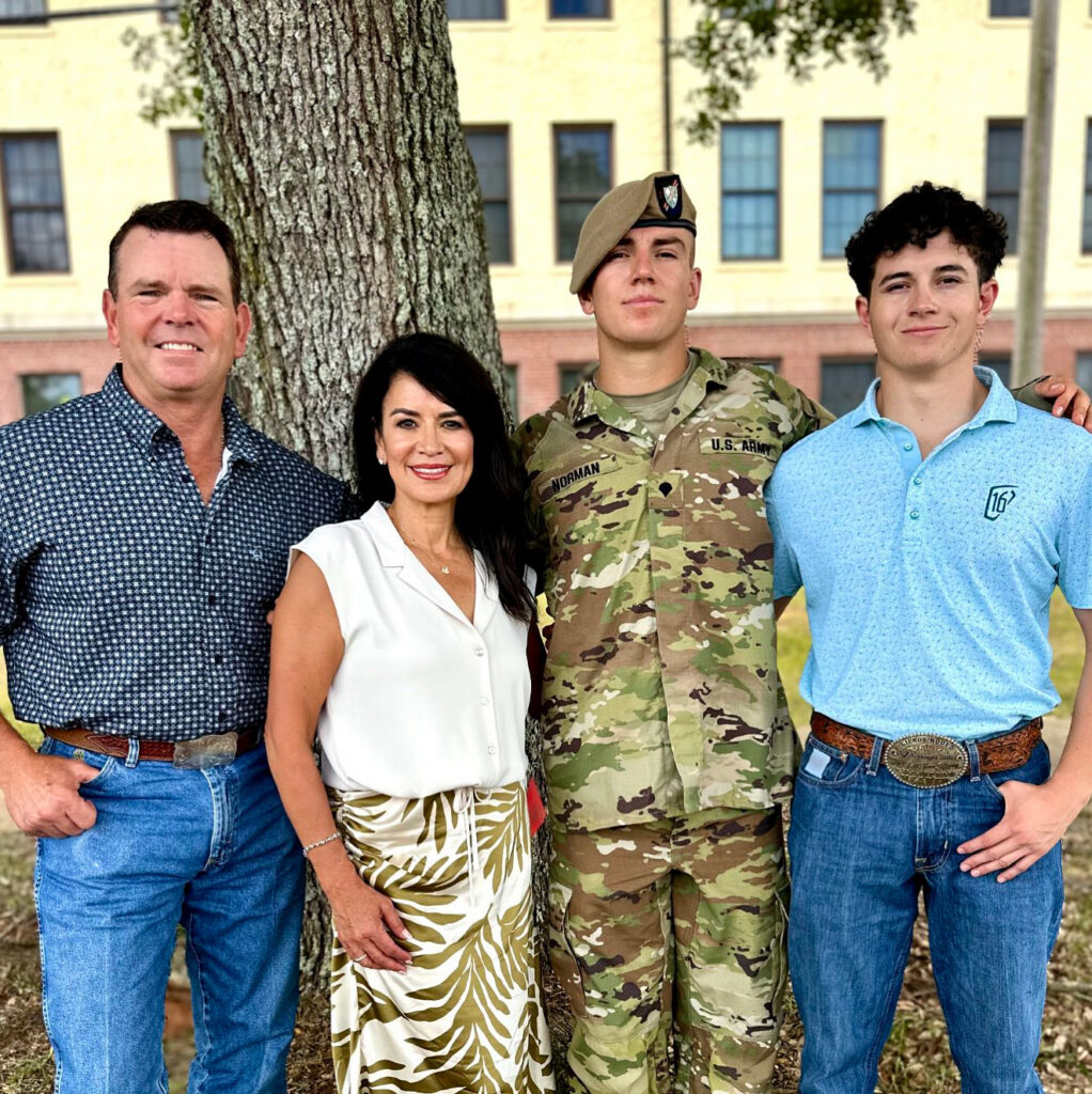 Bill Norman stands with his wife, DeAnna, and their two sons, one in a U.S. Army uniform and beret, in front of a tree and a brick building.