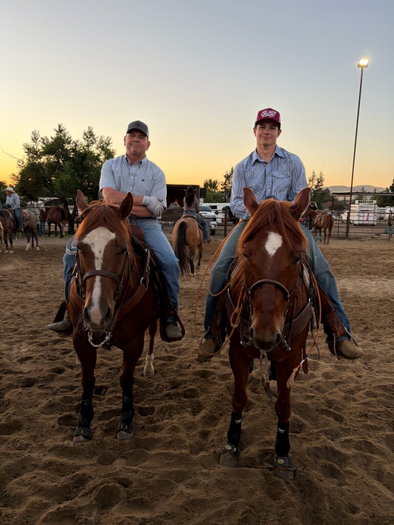 Bill Norman and one of his sons sit on sorrel horses with white blazes in a roping arena at sunset.