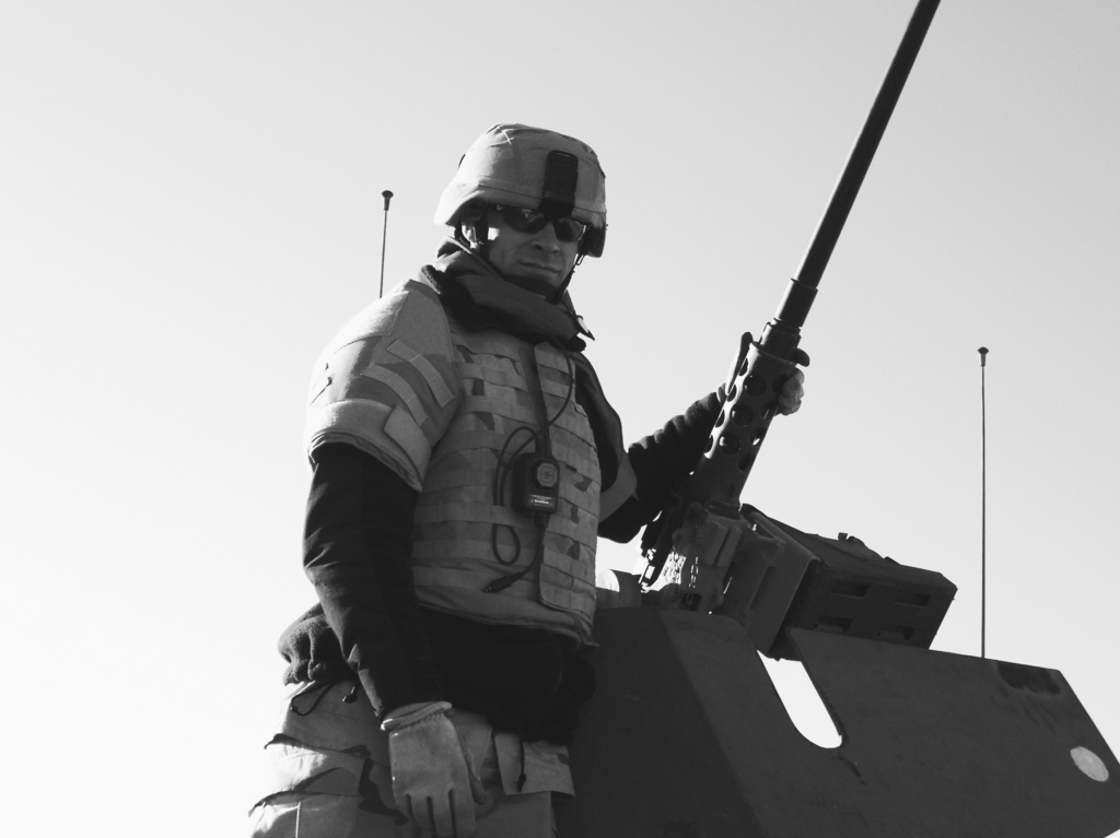 Jeremy Svejcar stands in a vehicle turret with a mounted machine gun in a black-and-white photo from his Army service.