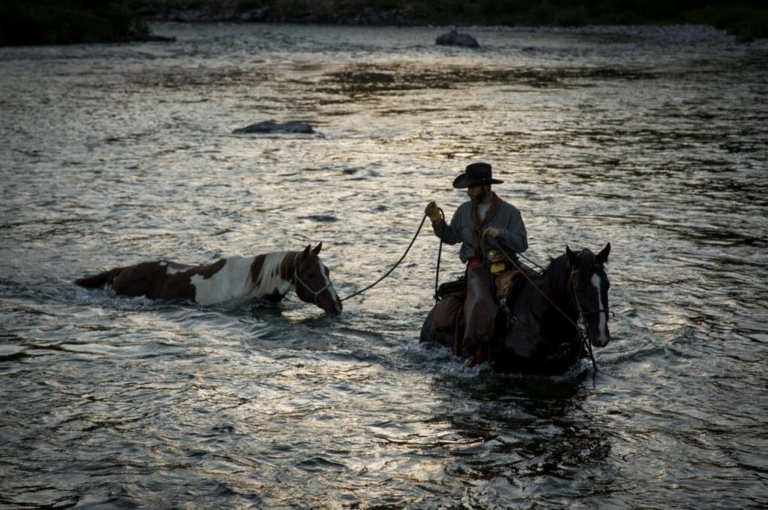 A cowboy on a dark horse ponies a brown-and-white paint horse across a shimmering river at dusk.