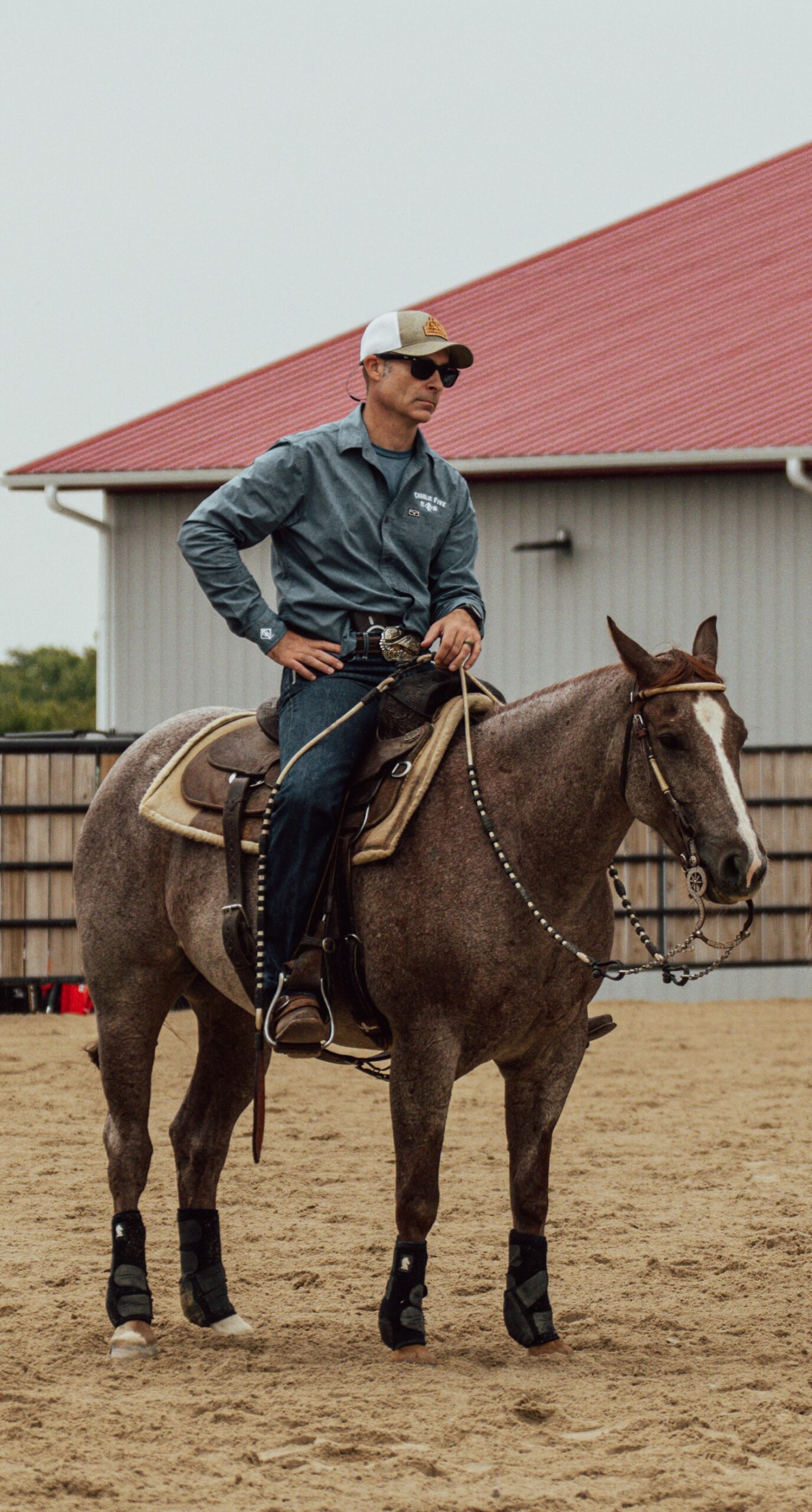 Jeremy Svejcar sits on a red roan horse in a sandy arena in front of a red-roofed barn.